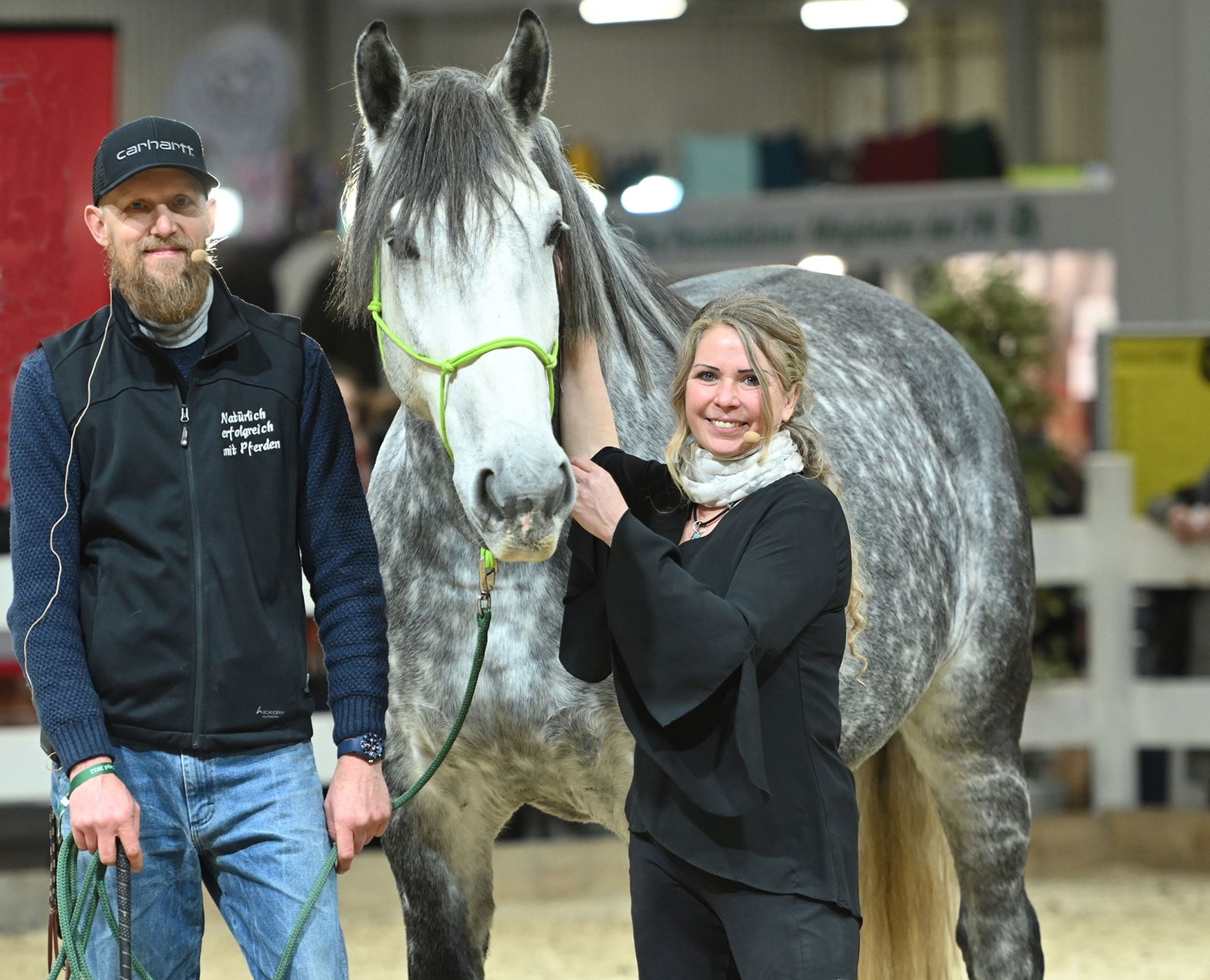 Das Bild zeigt zwei Personen, die neben einem großen, hellen Pferd stehen. Peer und Jenny sind auf der Messe PASSION PFERD 2025 zu sehen. Bei dem Pferd handelt sich um eine große Rasse mit einem gesprenkelten grauen Fell, einer markanten helleren grauen Mähne und einem markanten Schwanz. Es sieht gepflegt aus. Das Pferd trägt ein hellgrünes Halfter um den Kopf. Der Mann, Peer, ist mit einem Mikro ausgestattet, weil es sich auf dem Bild um eine der Veranstaltungen von Peer und Jenny handelt.