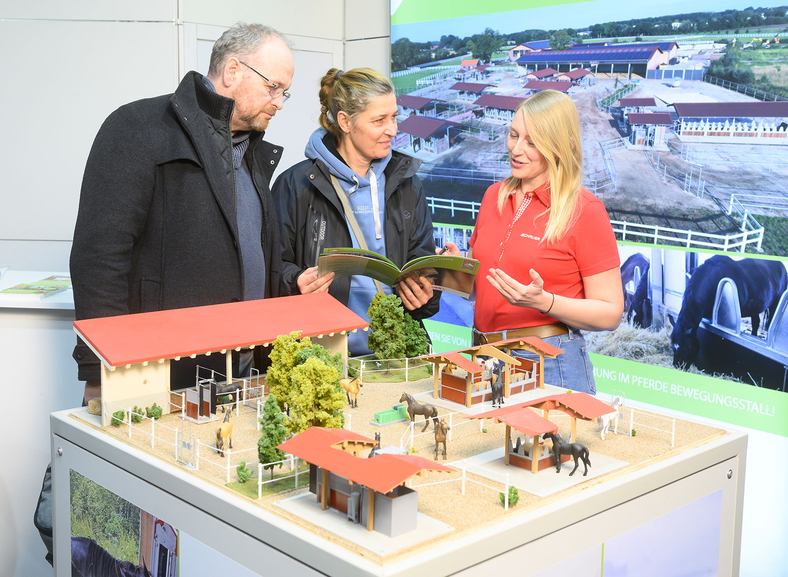Pferd.Zukunft.Stall Sonderschau auf der PASSION PFERD Hannover. Zwei Besucher lassen sich von einer Frau in einem roten Polohemd beraten. Im Vordergrund ist ein Modell einer Stallanlage zu sehen. 