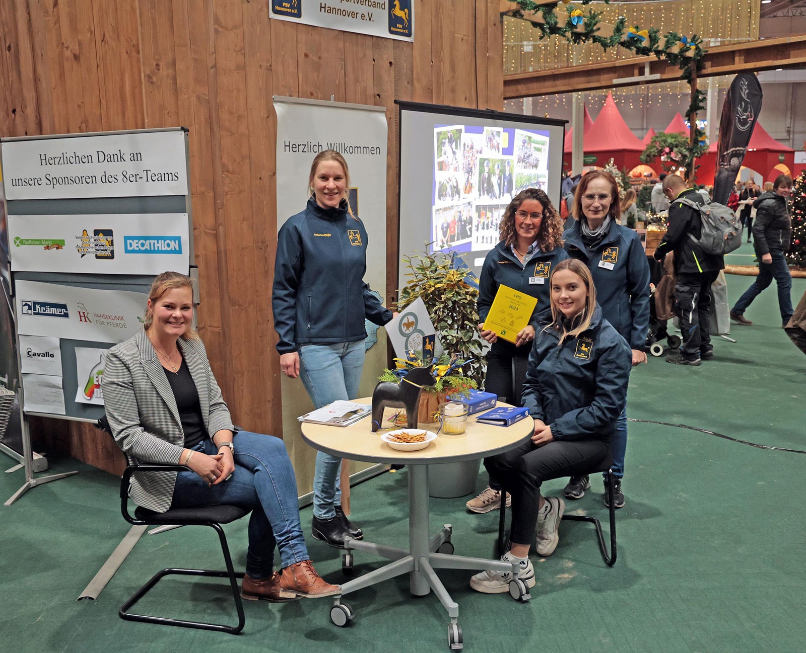 Das Bild zeigt eine Gruppe von fünf Personen vom Pferdesportverband Niedersachsen, die auf der Messe PASSION PFERD um einen Tisch an einem Stand stehen. Im Vordergrund befindet sich ein runder, heller Tisch mit vier Stühlen drumherum. Auf dem Tisch steht eine dunkle Pferdefigur, ein paar Broschüren und ein Stapel von Büchern oder Ordnern