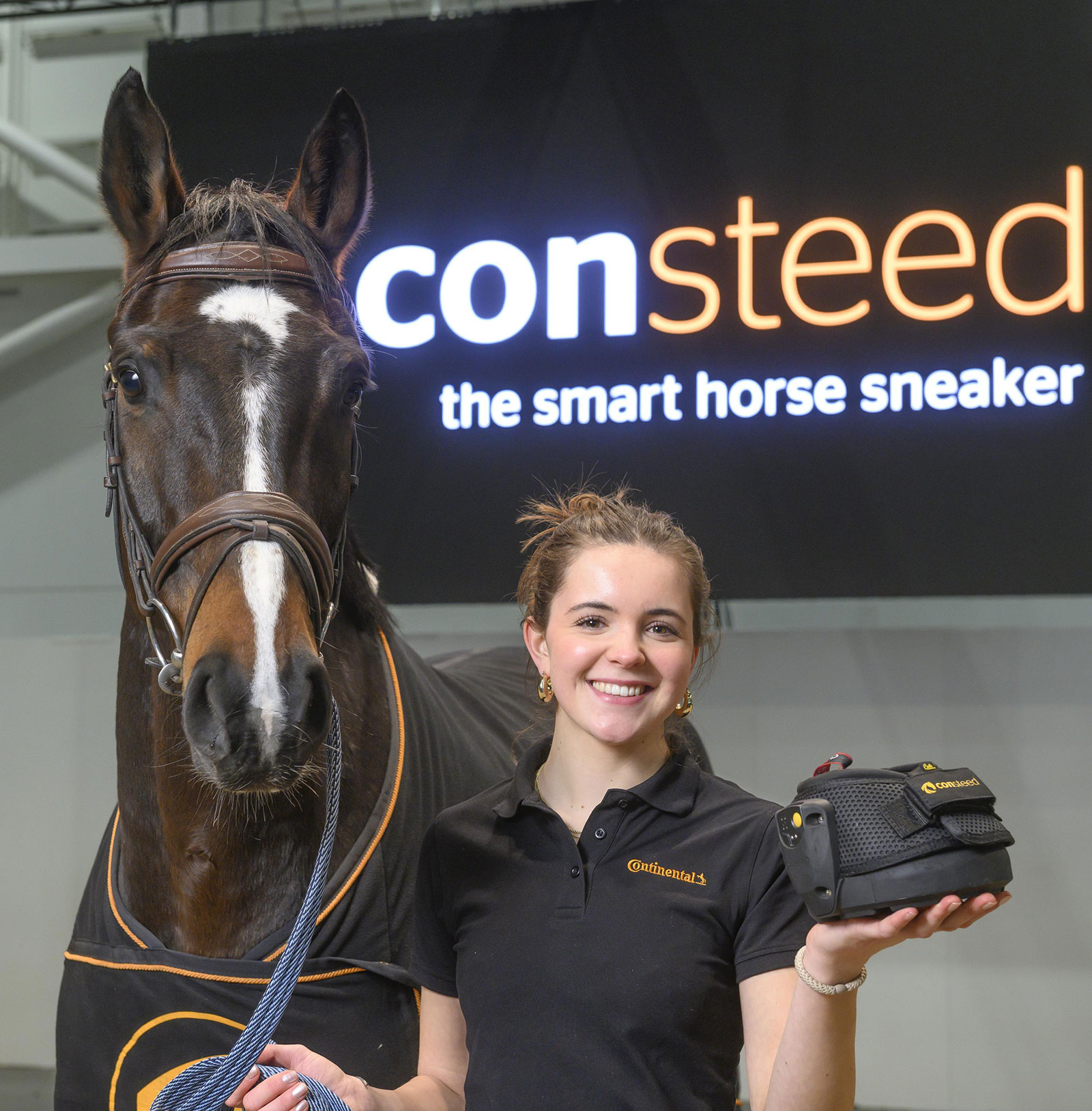 Werbebild, das eine junge Frau und ein braunes Pferd mit gepflegtem Fell zeigt, das eine dunkle Reitdecke über dem Körper trägt. Im Hintergrund befindet sich ein Leuchtschild mit der Aufschrift consteed, the smart horse sneaker. Die junge Frau trägt ein schwarzes Poloshirt mit dem Aufdruck der Firma Continental.
